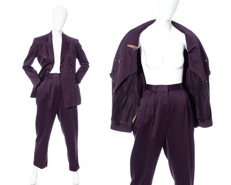 Pu&ograve; includere: Un completo viola con una giacca doppiopetto e pantaloni abbinati. La giacca ha una fodera marrone e i pantaloni hanno pieghe.