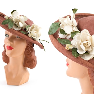 May include: A brown hat with white flowers and green leaves. The hat is worn by a mannequin with red lipstick.