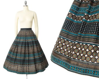 May include: A full-length skirt with a black, gold, and teal geometric pattern. The skirt is made of a thick, textured fabric and has a gathered waist.