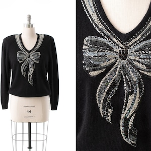 May include: Black sweater with a large, silver sequined bow on the front. The bow is made of silver and black sequins and has a V-neck.
