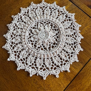 May include: A delicate, crocheted doily in a pale pink color. The doily features a circular design with intricate details and a raised center.