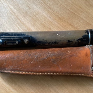 May include: A vintage cylindrical metal flashlight with a black and worn metallic finish, featuring a side-mounted switch and a rectangular lens, accompanied by a tan leather holster with a snap-button strap and a reinforced stitched edge.