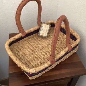 May include: A rectangular woven basket made of natural straw with two tan leather-wrapped handles. The basket features a dark purple and blue woven band near the rim and includes a rectangular tag labeled Bolga Baskets International.