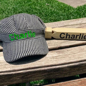 May include: A black and white striped engineer hat and a wooden whistle, both with the name "Charlie" printed on them in green and black, respectively. The hat is made of fabric and the whistle is made of wood.