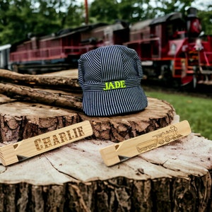 May include: Two wooden train whistles, one with the name "CHARLIE" engraved on it and the other with a train illustration. A blue and white striped engineer's cap with the name "JADE" embroidered on it sits on a tree stump.
