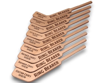 May include: Ten wooden hockey stick-shaped ring bearer signs with the names of the ring bearers and the date 10/12/18 engraved on each sign. The signs read "RING BEARER" in all caps.