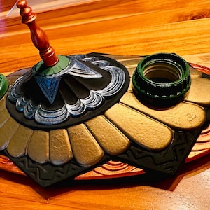 May include: A decorative, multi-piece object with a dark base, gold accents, and a central, ornate element. The piece includes a red and green top and two green rings. The object is on a wooden surface.