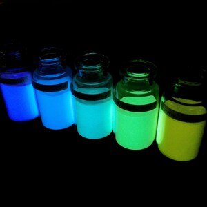May include: Five glass bottles filled with glowing liquid in different colors, including blue, green, and yellow. The bottles are arranged in a row on a dark surface.