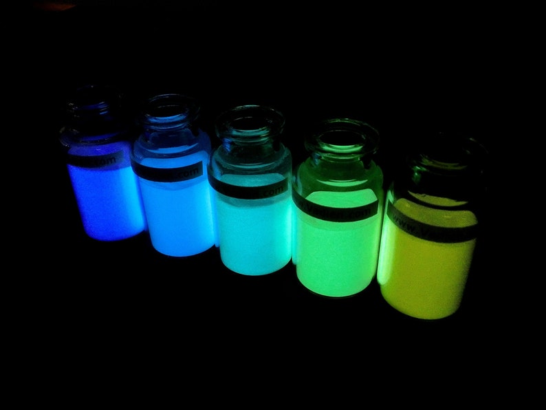May include: Five glass bottles filled with glowing liquid in different colors, including blue, green, and yellow. The bottles are arranged in a row on a dark surface.