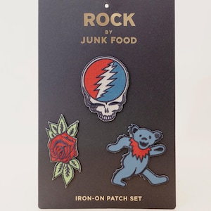 May include: A set of three iron-on patches on a black display card labeled ROCK BY JUNK FOOD. The patches include a red, white, and blue skull design, a red rose with green leaves, and a blue dancing bear with a red collar on a dark background.