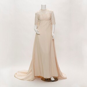 Puede incluir: Vestido de novia marfil vintage sobre un maniquí. El corpiño presenta un cuello alto y mangas cortas con delicados detalles de encaje. La falda fluye en una larga cola. El vestido se exhibe en un maniquí blanco sobre un fondo blanco.