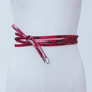 Può includere: Una cintura rossa e nera con motivo a pelle di serpente è avvolta attorno a un manichino bianco. La cintura è annodata sul davanti, con le estremità che pendono. La cintura ha puntali metallici color argento.