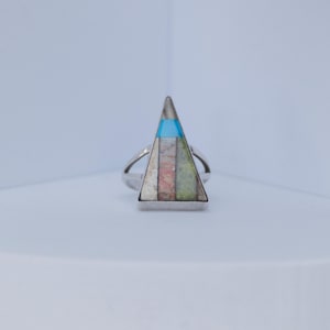 Op de afbeelding: Een zilveren ring met een driehoekige inleg. De inleg heeft een geometrisch patroon met blauwe, roze, groene en witte secties. De ring staat tegen een witte achtergrond.