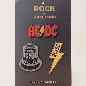 Peut inclure: Un ensemble de trois écussons thermocollants sur une carte noire. Les écussons présentent le logo et les images du groupe AC/DC, dont un logo rouge et jaune, un éclair jaune et une cloche avec les mots "Hells Bells". La carte indique "ROCK by JUNK FOOD".