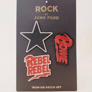 Puede incluir: Un conjunto de cuatro parches termoadhesivos sobre una tarjeta gris oscuro. Los parches incluyen una estrella negra con ribete blanco, un parche rojo y negro con la palabra "BOWIE" y un parche rojo y blanco que dice "REBEL REBEL". La tarjeta dice "ROCK BY JUNK FOOD".