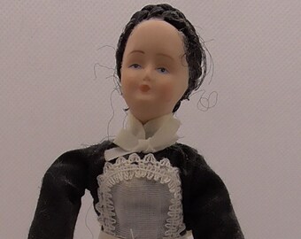 May include: A porcelain doll wearing a black dress with a white apron and lace trim. The doll has black hair and a painted face with blue eyes.