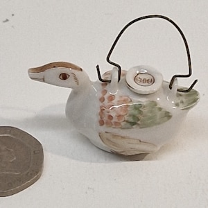 Puede incluir: Pequeña figura de pato de porcelana blanca, con pico y cabeza marrones, y un asa de alambre. El pato tiene detalles rosas y verdes en el cuerpo y una pequeña tapa redonda con texto japonés.