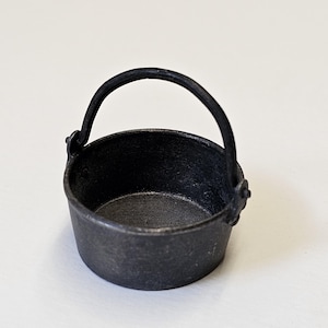 Puede incluir: Una pequeña olla de hierro fundido negra con asa. La olla tiene un diámetro de unos 10 cm y una altura de 7,5 cm.