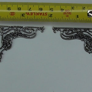 Puede incluir: Dos soportes de esquina de metal plateado con un diseño floral decorativo. Los soportes miden aproximadamente 5 cm de largo.