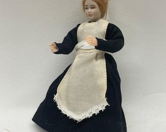 May include: A miniature doll dressed in a traditional black and white outfit, including a long black skirt, a white apron with lace trim, and a white bonnet. The doll has red hair and is posed with arms outstretched.
