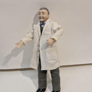 May include: A miniature doll figure with grey hair and a moustache, wearing a white lab coat over a grey vest and trousers. The figure has a blue patterned bowtie, a light-coloured shirt, and black shoes. The doll is posed with arms extended outwards.