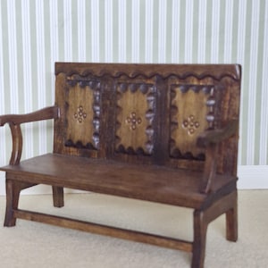 May include: A miniature wooden bench with a dark brown finish. The back of the bench features three rectangular panels with carved floral designs. The bench has armrests and a scalloped top edge, perfect for a dollhouse or miniature scene.