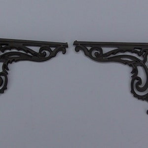 May include: Two black metal corner brackets with a decorative, ornate design. The brackets are made of a thin, delicate metal and have a slightly distressed finish.