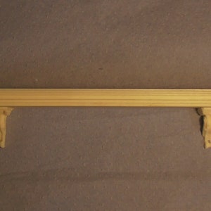 Può includere: Una mensola in legno con mensole decorative. La mensola è incompiuta, mostrando la venatura naturale del legno. Le mensole sono intagliate in modo intricato, aggiungendo un tocco classico. La mensola è lunga circa 122 cm.