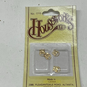 May include: A packet of gold-coloured decorative embellishments. The packet has the text "Houseworks LTD" and "Made in China". The embellishments are in a clear plastic container. The embellishments have a floral design.