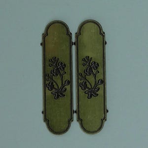 May include: Two connected, olive-green metal panels with a floral design. The panels have a rectangular shape with a curved top and bottom. The floral design is dark brown and is centered on each panel.