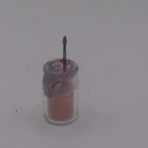 Puede incluir: Un pequeño frasco de vidrio con una tapa roja y blanca y una varilla de metal que sobresale de la parte superior. El frasco está lleno de un polvo naranja claro.