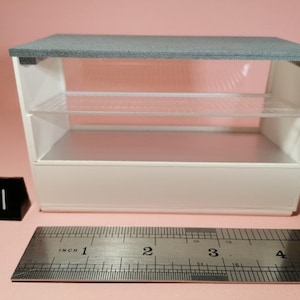 Puede incluir: Una vitrina blanca con una tapa gris y dos estantes de vidrio. La vitrina mide aproximadamente 30 cm de ancho y 25 cm de alto.