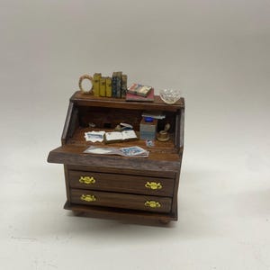 Puede incluir: Pequeño escritorio de madera oscura con una superficie de escritura abatible y dos cajones. El escritorio es de color marrón oscuro con tiradores dorados. La estantería superior contiene libros en miniatura, un pequeño cuenco de cristal y otros objetos decorativos.