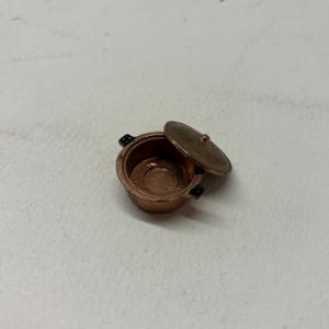 Può includere: Una piccola pentola color rame con coperchio leggermente socchiuso. La piccola pentola ha due manici neri e un coperchio abbinato. L'interno della pentola è visibile, mostrando una base circolare. Questo articolo è probabilmente decorativo.