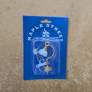 May include: A miniature gold lamp with a white lampshade, packaged in a blue card with the text "MAPLE STREET 12V Lighting Systems". The lamp has a curved arm and a round base, suitable for dollhouses or model displays.