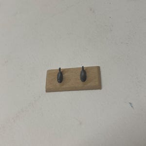 May include: A small, rectangular wooden wall hook with two dark gray metal hooks. The wood has a natural, light brown color. The hooks are evenly spaced and appear to be for hanging items.