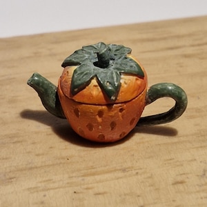 Puede incluir: Una pequeña tetera de cerámica con forma de fresa. La tetera es naranja con hojas verdes, un pico verde y un asa verde. Un objeto decorativo.