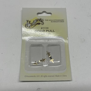 Puede incluir: Un paquete de tiradores de puerta Houseworks. El paquete incluye dos tiradores de puerta en miniatura de color dorado. El paquete tiene el texto "#1110 DOOR PULL 1" y "THE GOLD STANDARD IN MINIATURES".
