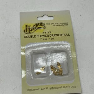 May include: A packet of gold-coloured double flower drawer pulls for miniature projects. The package contains four pieces, each with a delicate floral design. The label reads "Double Flower Drawer Pull" and "1" Scale".