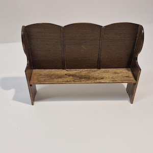 May include: A miniature wooden bench with a dark brown finish. The bench has a three-panel back and a simple seat. The wood grain is visible, adding texture. The bench is designed for a dollhouse or miniature setting.