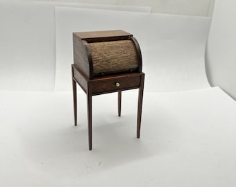May include: Miniature wooden roll-top desk with a small drawer and four slender legs. The desk features a curved, slatted roll-top cover and a small brass knob on the drawer. The desk is made of dark wood and is a vintage style.