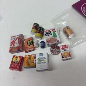 Könnte beinhalten: Miniatur-Lebensmittel, darunter Müslipackungen, Obstkonserven, eine Rosinenbox und eine Zuckerdose. Die Kellogg's Corn Flakes-Box zeigt einen Hahn. Die Ritz-Box zeigt Cracker. Die Just Juice-Box zeigt eine Orange.