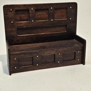 Può includere: Una mensola da parete in legno marrone scuro con un pannello posteriore e un ripiano inferiore. Il pannello posteriore ha tre sezioni rettangolari incassate ed è adornato da piccoli borchie color ottone. Il ripiano inferiore ha anche sezioni incassate e borchie.