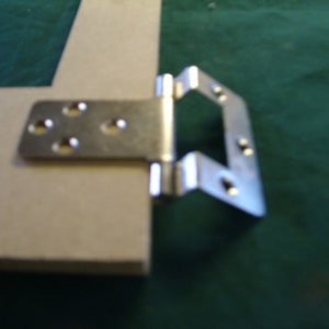 Puede incluir: Bisagra de metal plateado con varios agujeros para tornillos. La bisagra está unida a una pieza de madera.