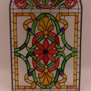 Peut inclure: Un panneau de vitrail avec un motif floral. Le panneau pr&eacute;sente un motif floral rouge, jaune et vert avec un centre bleu. Le panneau est encadr&eacute; d'un cadre en m&eacute;tal noir.