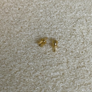 May include: Two small, gold-coloured metal electrical connectors. One connector has a cylindrical shape with a small opening, and the other has a rectangular shape with a small opening. The connectors are on a textured, light-coloured surface.