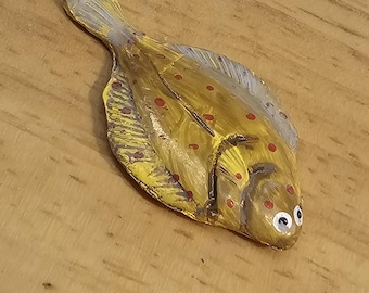 Puede incluir: Un pez de madera pintado de amarillo y gris con manchas rojas y ojos blancos. El pez está acostado sobre una superficie de madera.