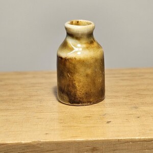 Può includere: Un piccolo vaso in ceramica marrone con un bordo bianco. Il vaso ha una smaltatura marrone macchiata.