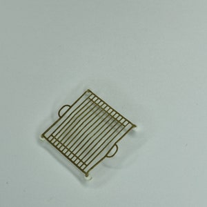 Peut inclure: Une grille de refroidissement en métal de couleur dorée de forme carrée avec des coins arrondis. La grille a une forme de grille et quatre petits pieds blancs.