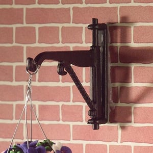 Puede incluir: Un soporte colgante de metal negro ornamentado, montado en una pared de ladrillos rojos. El soporte tiene un brazo curvado y un soporte decorativo, diseñado para sostener una cesta colgante. Se ve una cadena plateada y una cesta colgante con flores moradas y blancas.